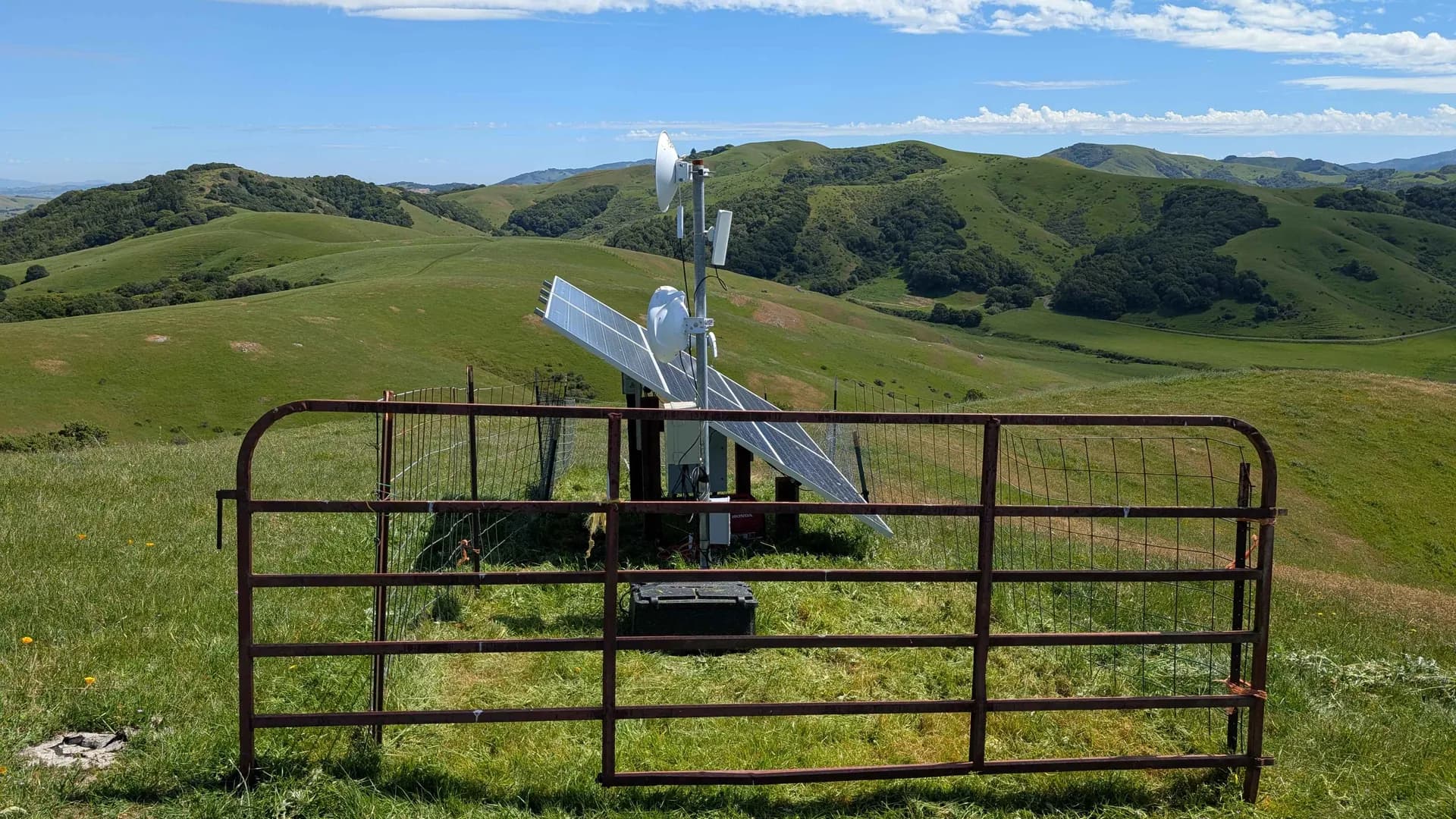 Rust-orange ranch gate framing an off-grid solar relay with rolling emerald Sonoma hills behind.